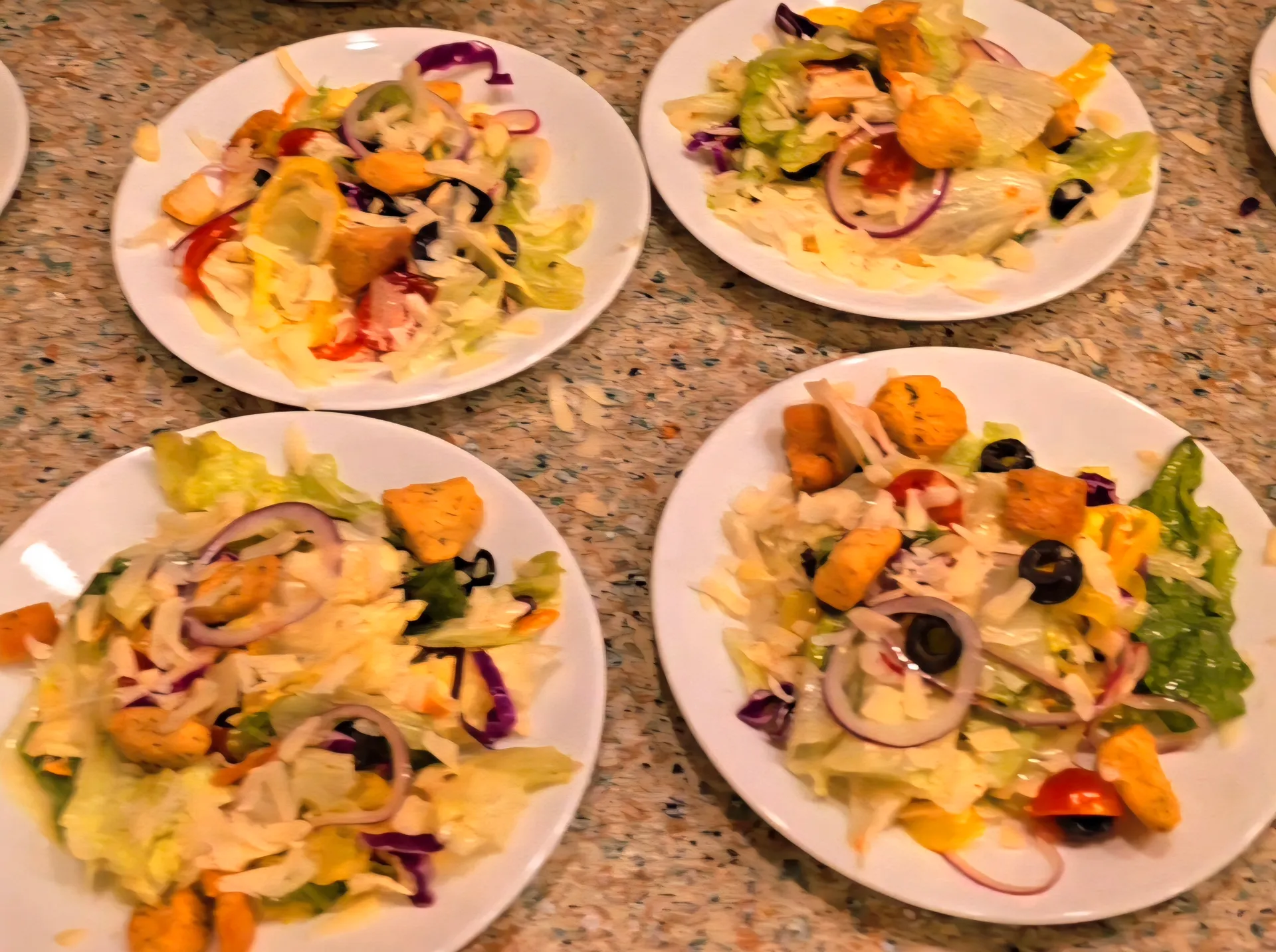 Four white plates with fresh salads featuring mixed greens, sliced peppers, olives, and croutons on granite counter