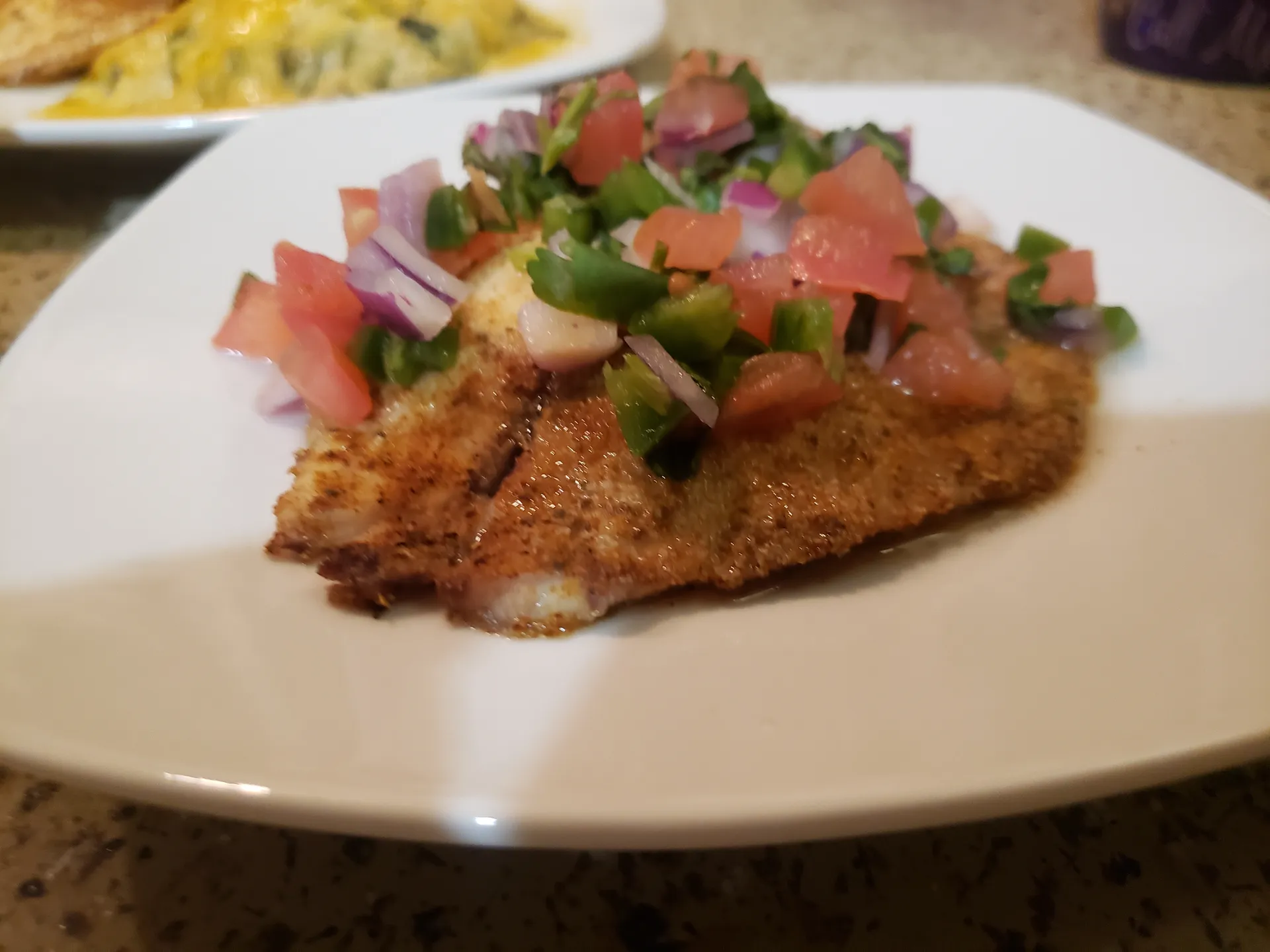 Breaded fish fillet topped with fresh pico de gallo salsa with diced tomatoes, onions, and peppers on white plate