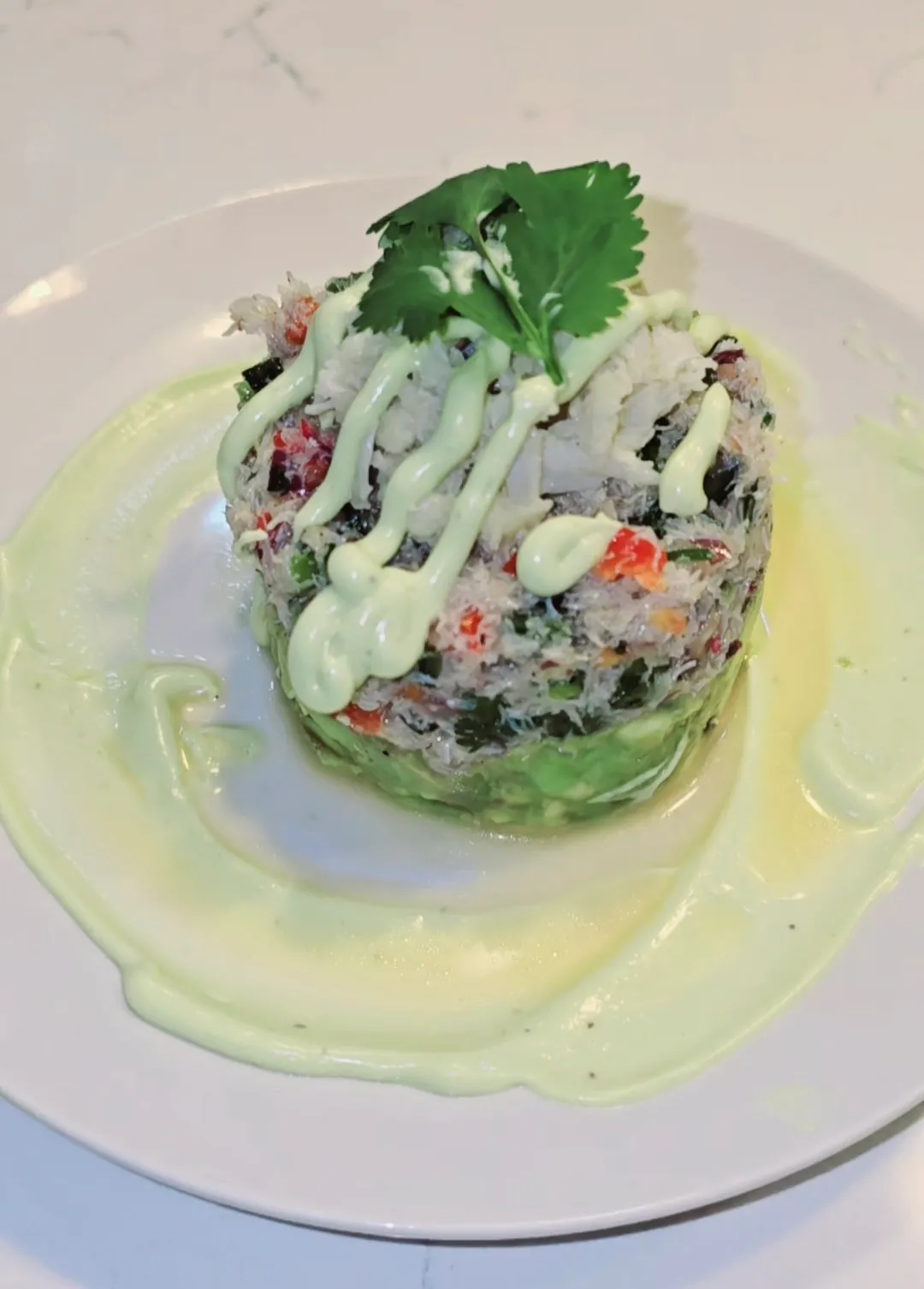 Layered crab salad tower with avocado base, drizzled with sauce and garnished with cilantro on white plate