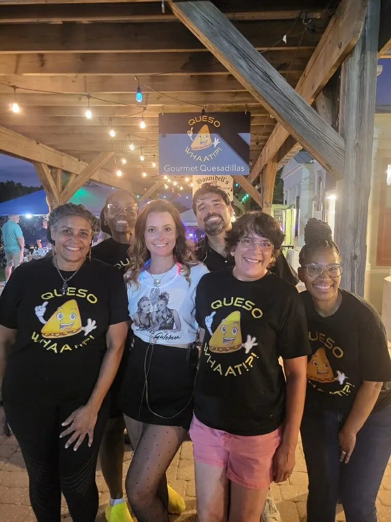 Six team members in QuesoWhaat branded shirts posing together at their outdoor food booth under string lights