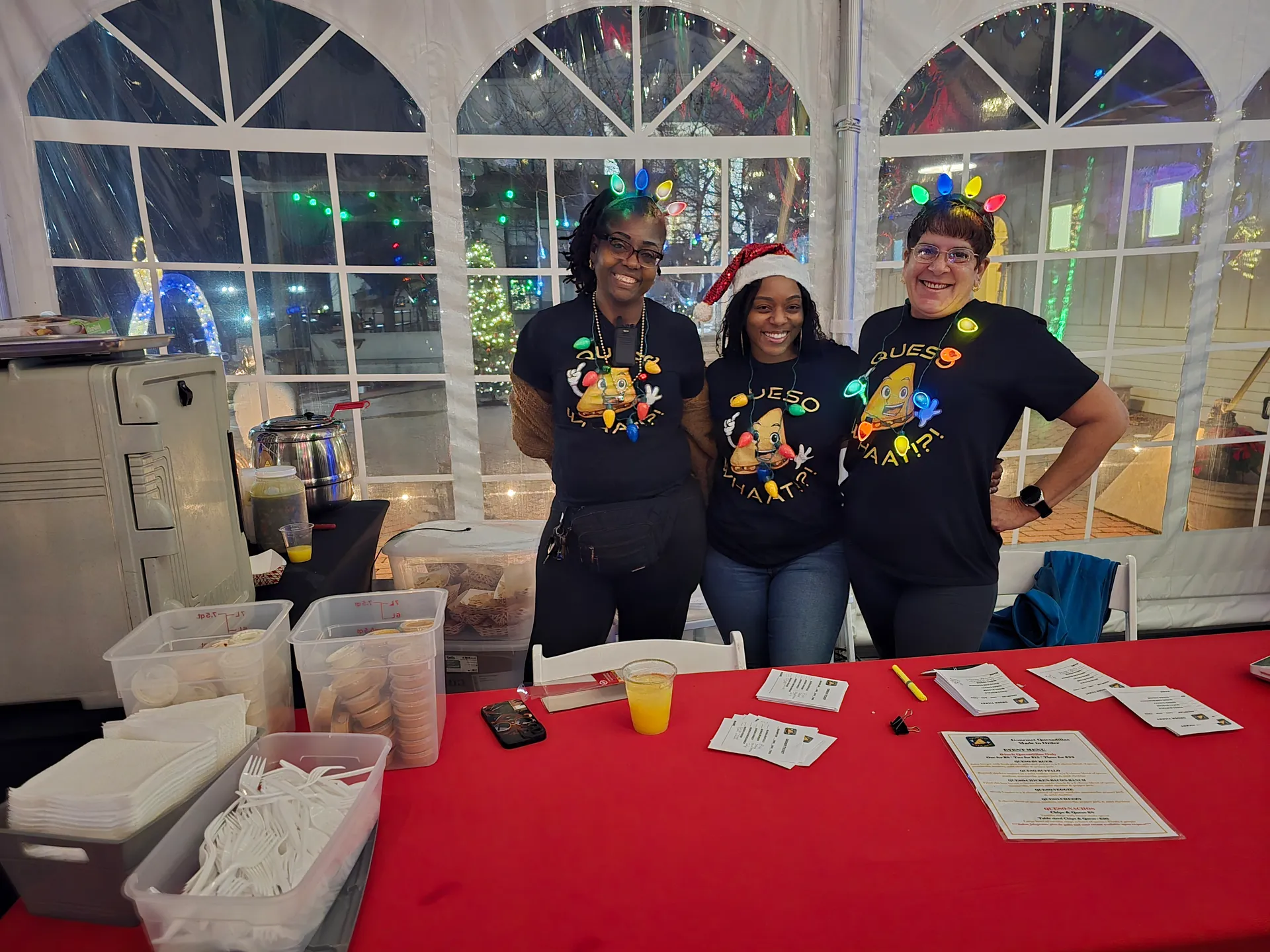 Three smiling staff members wearing matching QuesoWhaat!?! t-shirts and festive headbands behind a catering table