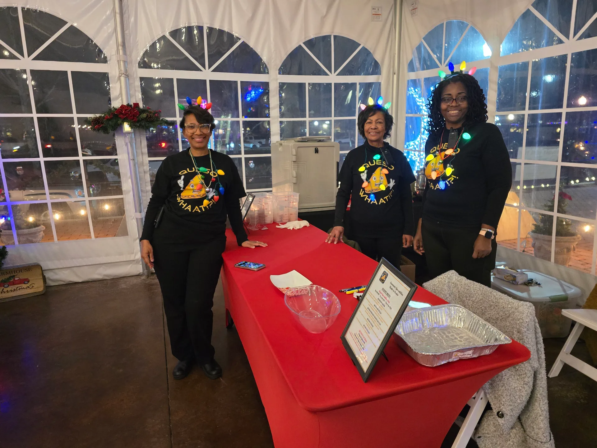Three women in matching black Whaat!?! hoodies with festive headbands behind red-draped catering tables at event