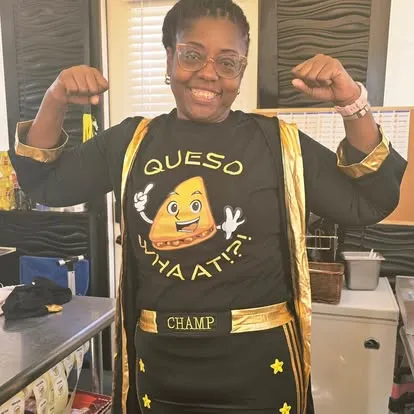 Chef in black shirt with queso graphic and gold accents, flexing arms in kitchen setting