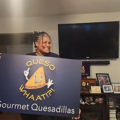 Smiling person holding purple Queso Whaat!?! Gourmet Quesadillas banner with cartoon cheese logo in home setting