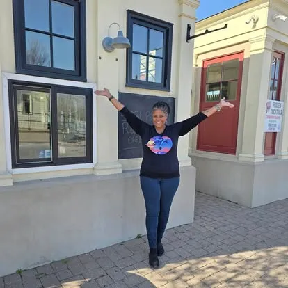 Woman in black outfit with tie-dye heart logo standing with arms outstretched in front of modern building exterior