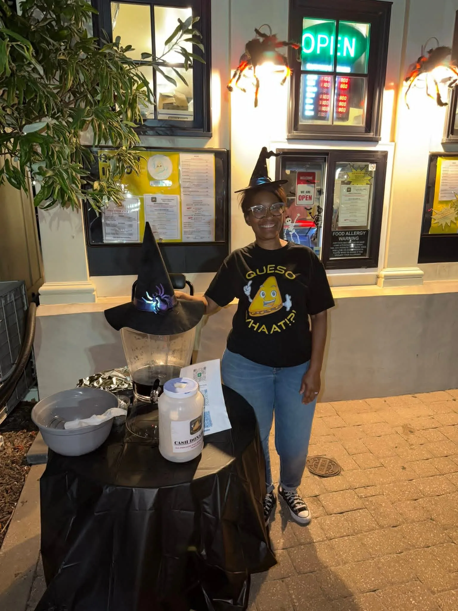Smiling team member in witch hat holding decorated witch hat prop at Whaat!?! Foods restaurant counter