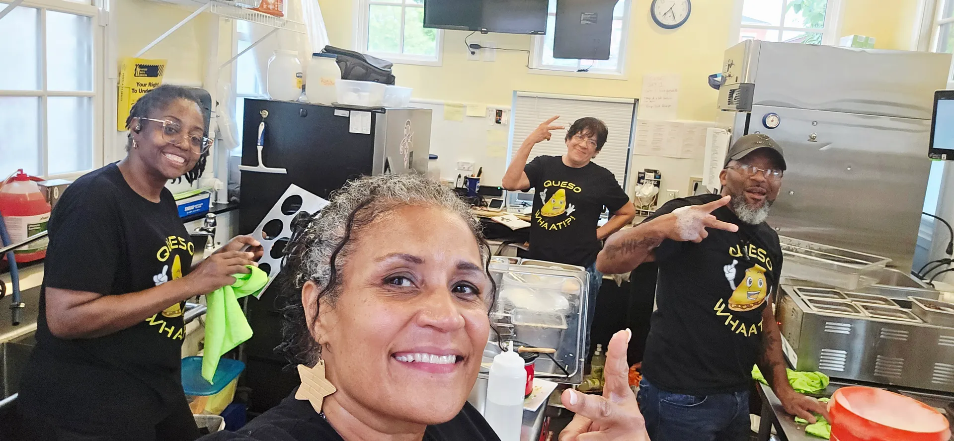 Four team members in black Queso Whaat t-shirts smiling and posing in commercial kitchen with stainless steel equipment
