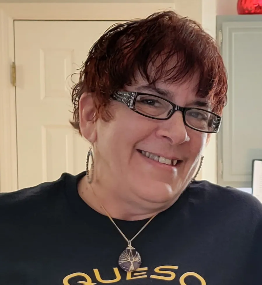 Smiling woman with glasses and short reddish hair wearing a black QuesoWhaat shirt with pendant necklace