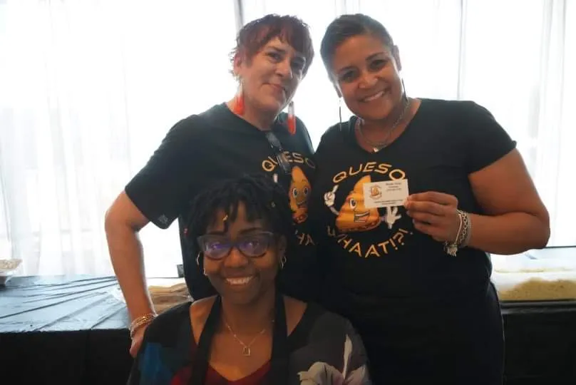 Three team members wearing black QuesoWhaat!?! branded t-shirts smiling together, one holding a business card